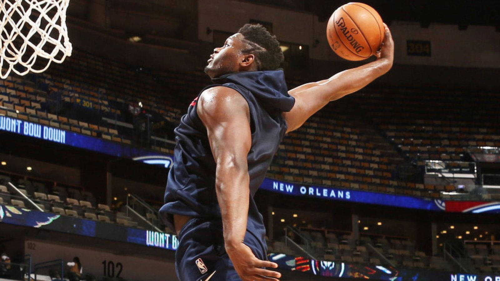 錫安·威廉森 NBA訓練 壁紙 — 一名身穿海軍藍球衣的籃球運動員在一個帶有'New Orleans'標牌和空座位的場館中扣入Spalding籃球。