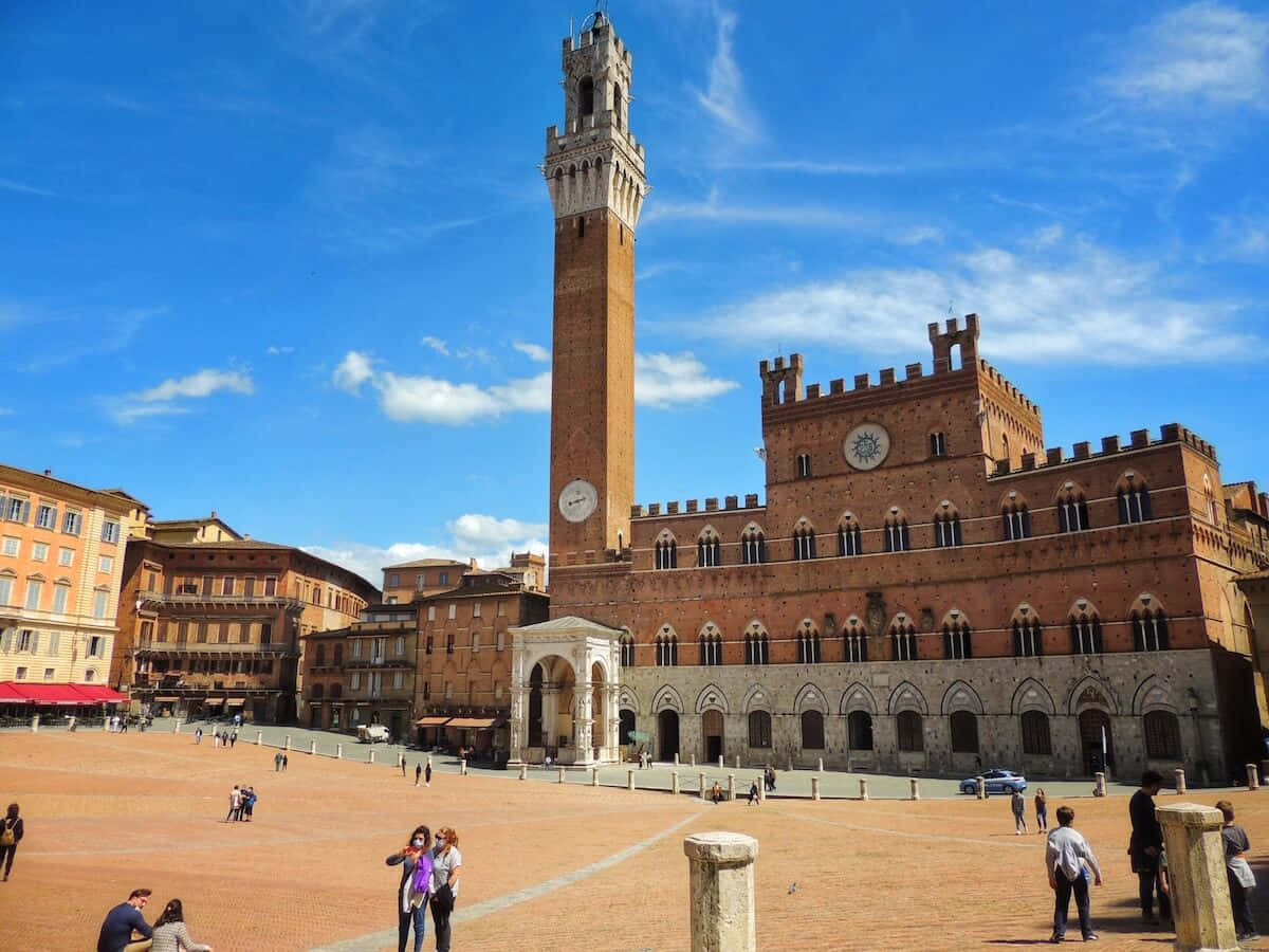 錫耶納公眾宮殿 壁紙 — 在Piazza del Campo陽光明媚的一天，標誌性的Torre del Mangia和Palazzo Pubblico聳立在陶土廣場上的遊客之上。