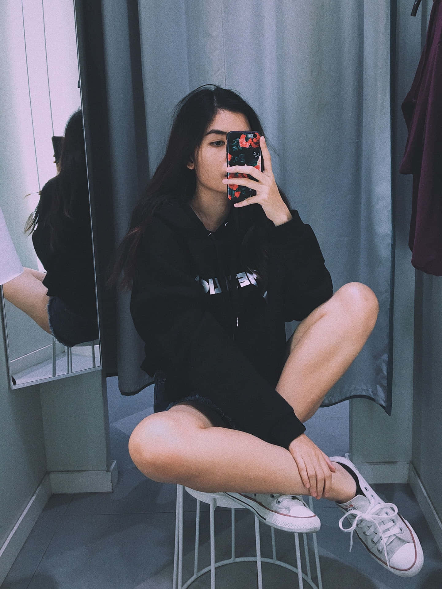 年輕女性在試衣間對著鏡子自拍，身穿黑色連帽衫和白色運動鞋。