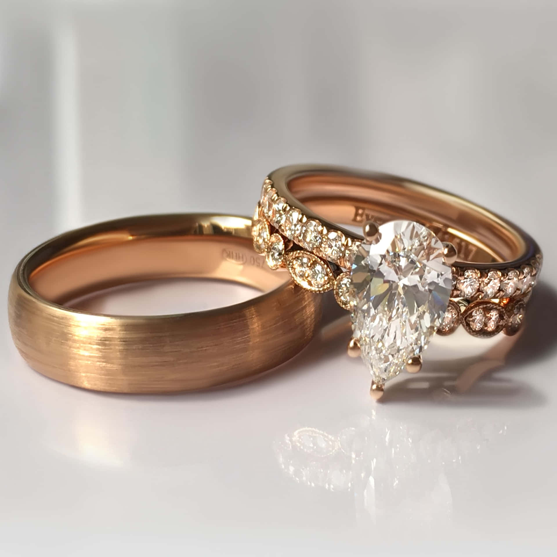 兩枚帶有鑽石的玫瑰金結婚戒指放在白色反光表面上。