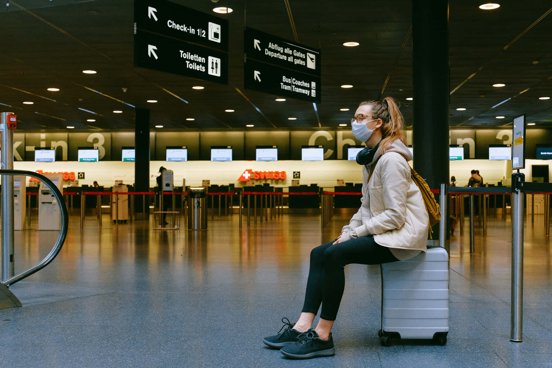 一名戴著口罩的旅客坐在一個空蕩蕩的機場客運大樓內的行李箱上。