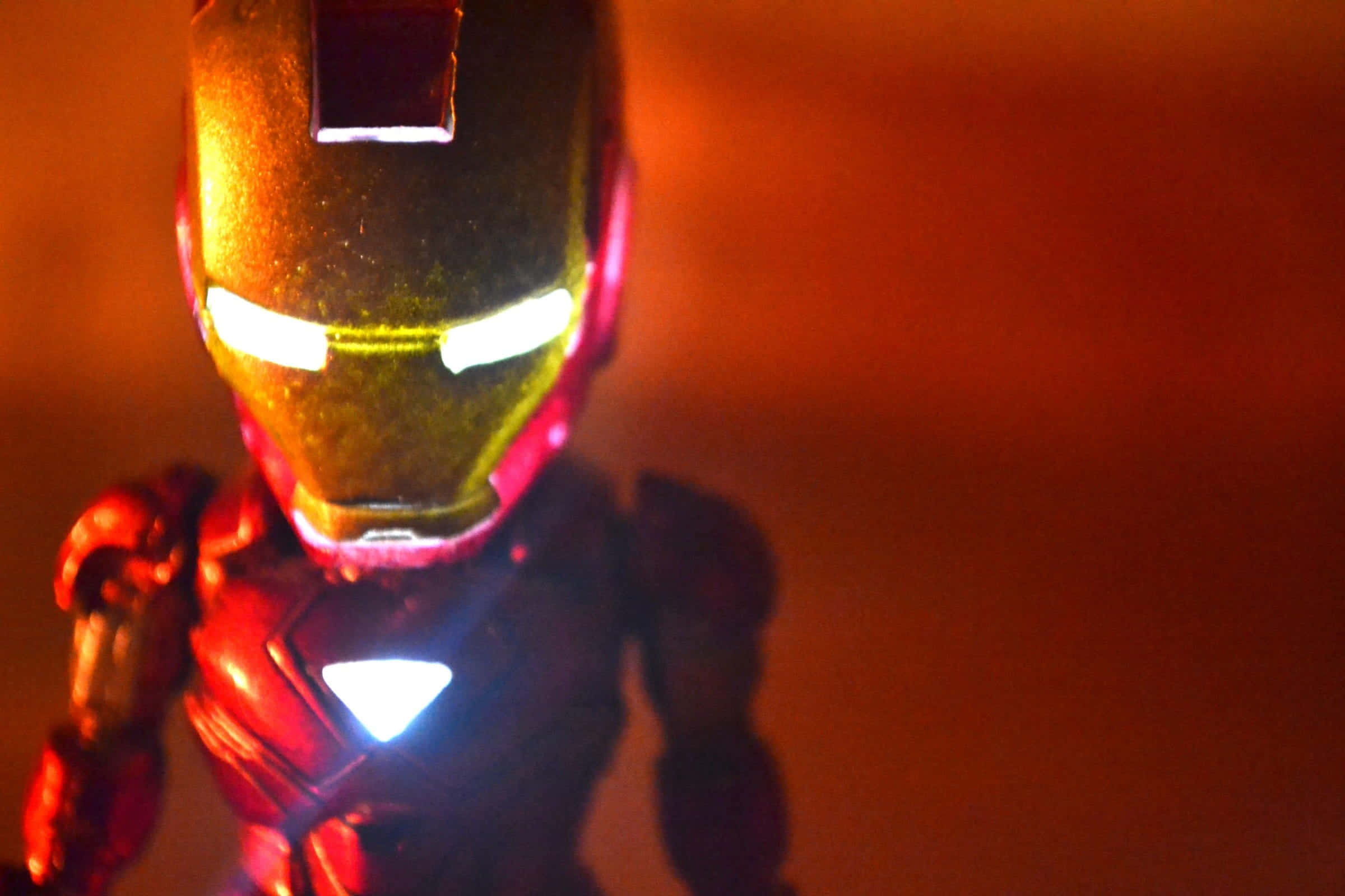 Iron Man Action Figures 桌布 壁紙 — 鐵甲奇俠動作人偶的特寫，眼睛和胸部弧形反應器發光，背景為溫暖的橙色。