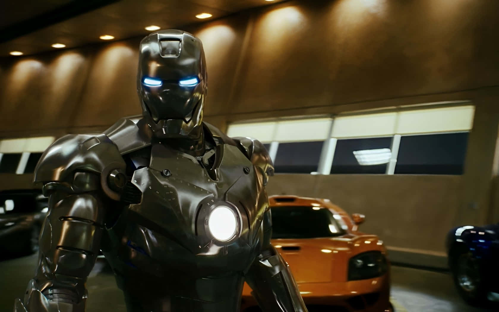 Iron Man Suit 桌布 壁紙 — 鐵甲奇俠站在時尚的車庫中，眼睛和胸部弧形反應堆發光，旁邊是一輛充滿活力的橙色跑車。
