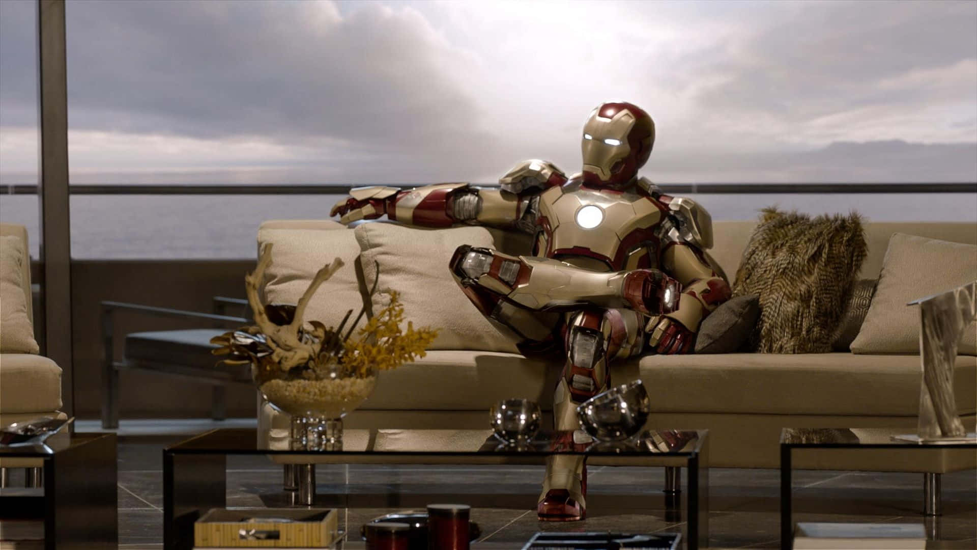 Iron Man Movies 桌布 壁紙 — 鐵甲奇俠身穿紅金色盔甲，在雲層密佈的天空下，於一間可俯瞰海洋的現代玻璃牆頂層公寓中，隨意地躺在一張米色沙發上。