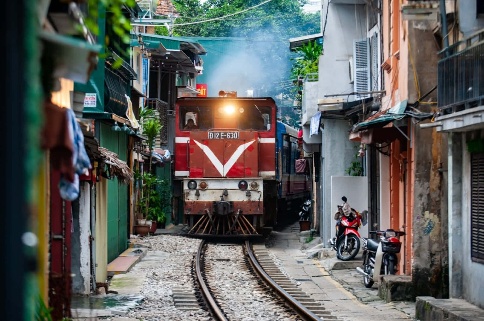 紅色火車在狹窄的都市小巷中行駛，旁邊有電單車和色彩繽紛的建築物。