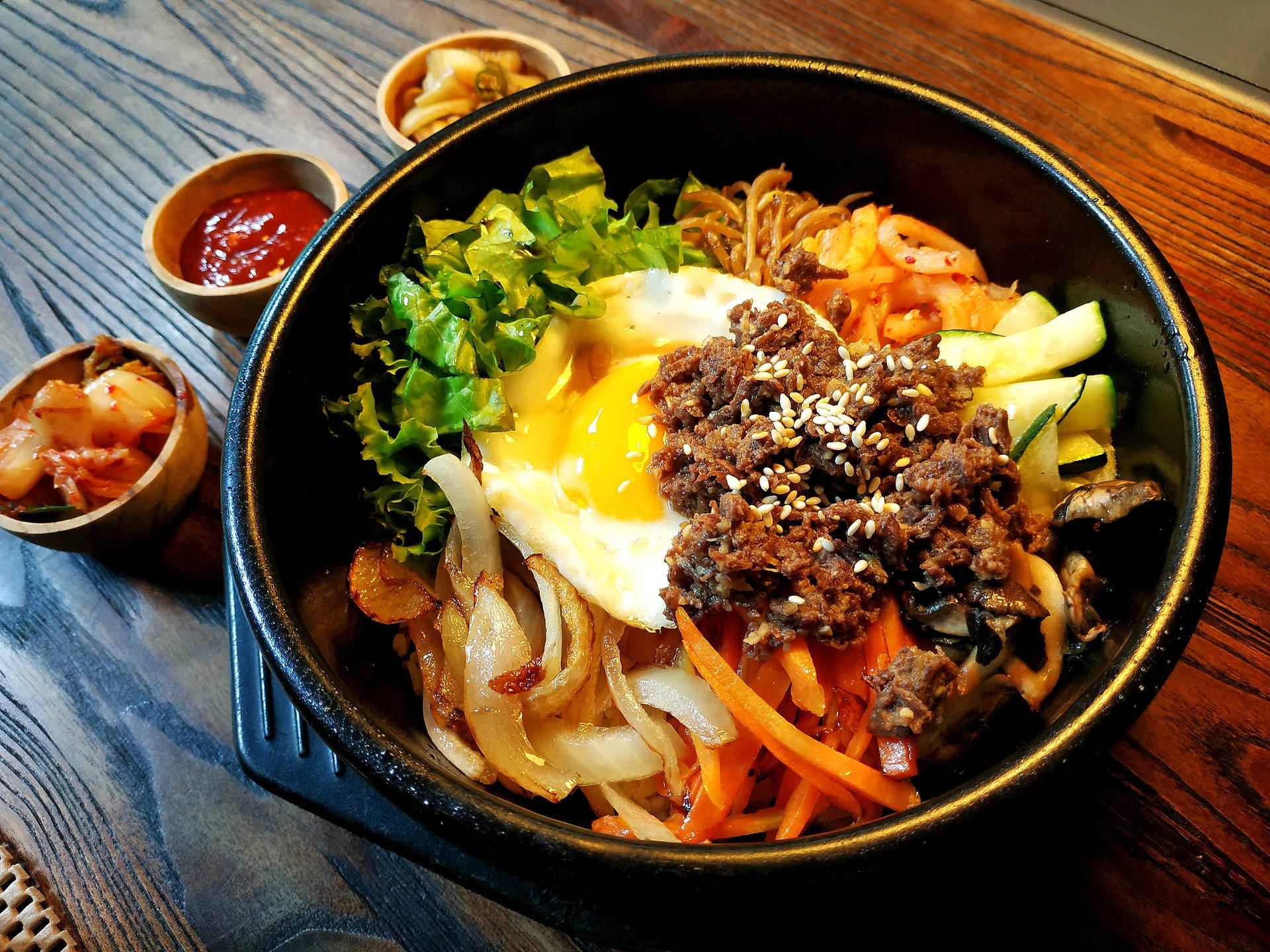 鑄鐵碗盛裝的韓式拌飯 壁紙 — 一個冒煙的石鍋拌飯，上面有太陽蛋、芝麻和色彩繽紛的蔬菜，放在質樸的木桌上。