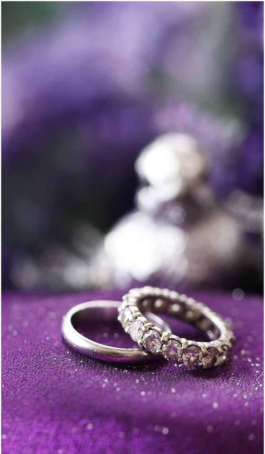 兩枚結婚戒指放在紫色閃粉布料上，背景有模糊的花朵。