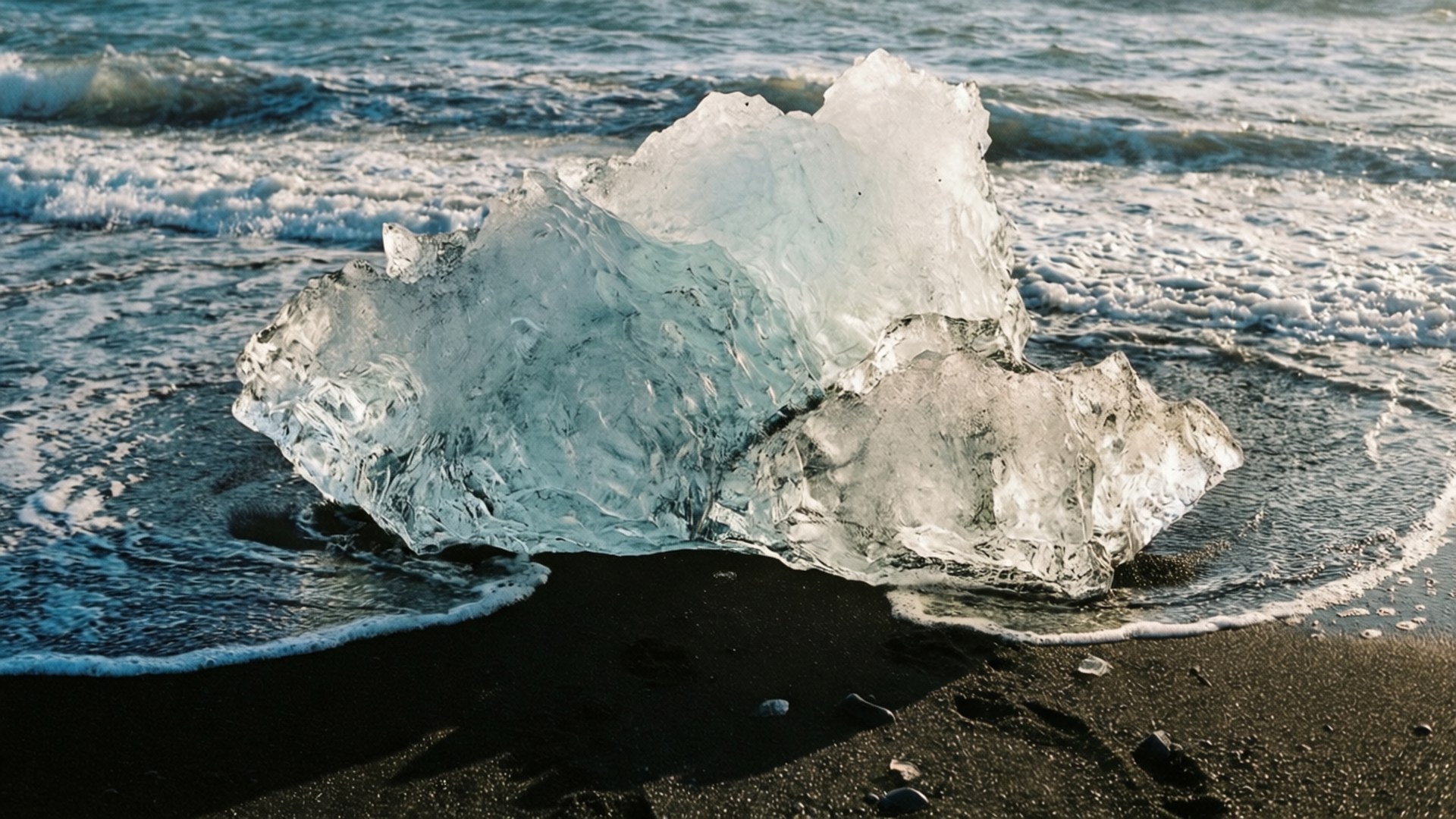 鑽石沙灘 壁紙 — 一座巨大的半透明冰山坐落於黑色火山海灘上，溫柔的海浪拍打著其晶瑩的底部。