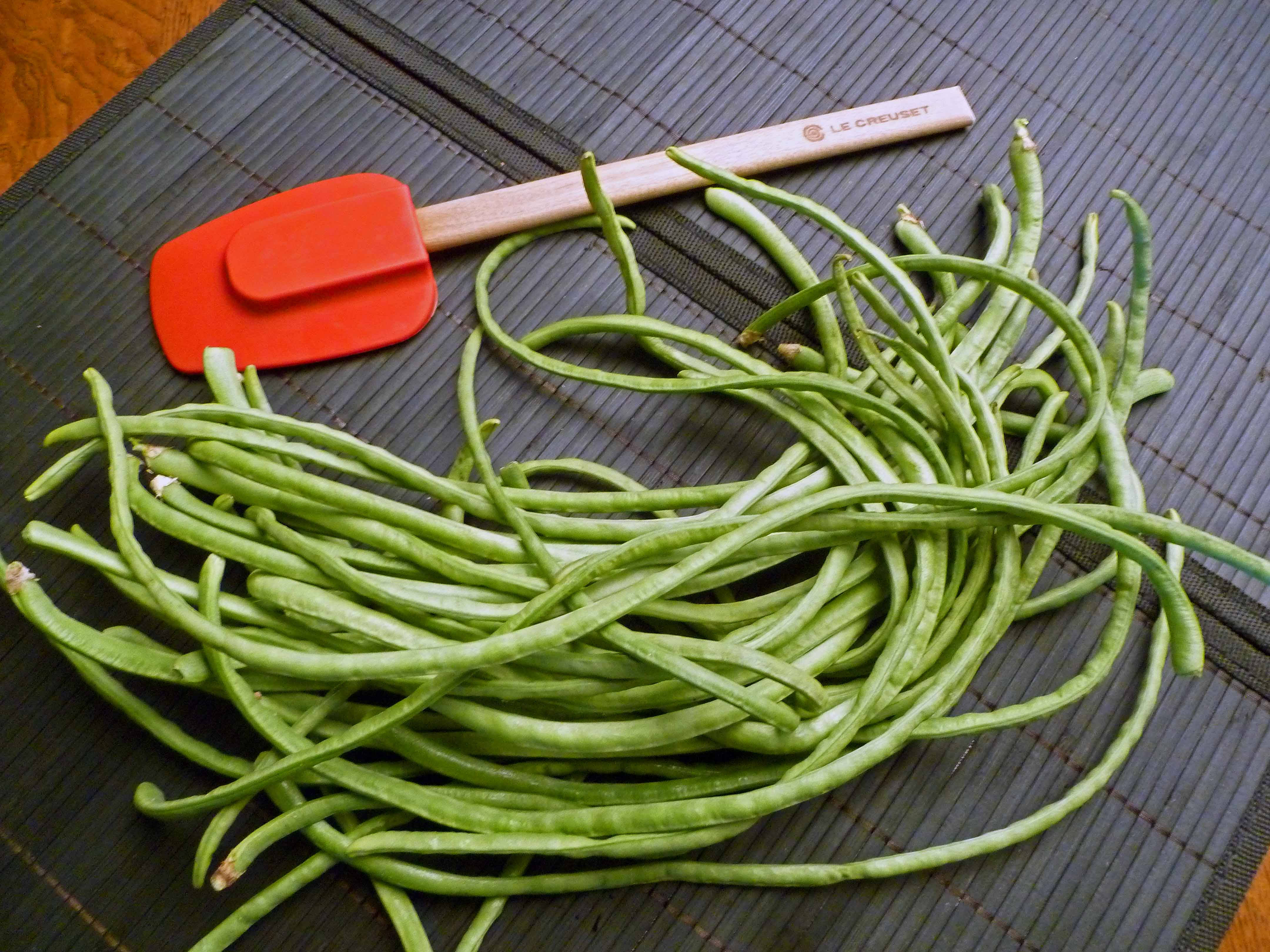 長四季豆 壁紙 — 新鮮青豆堆在深色竹蓆上，旁邊有紅色矽膠鍋鏟。