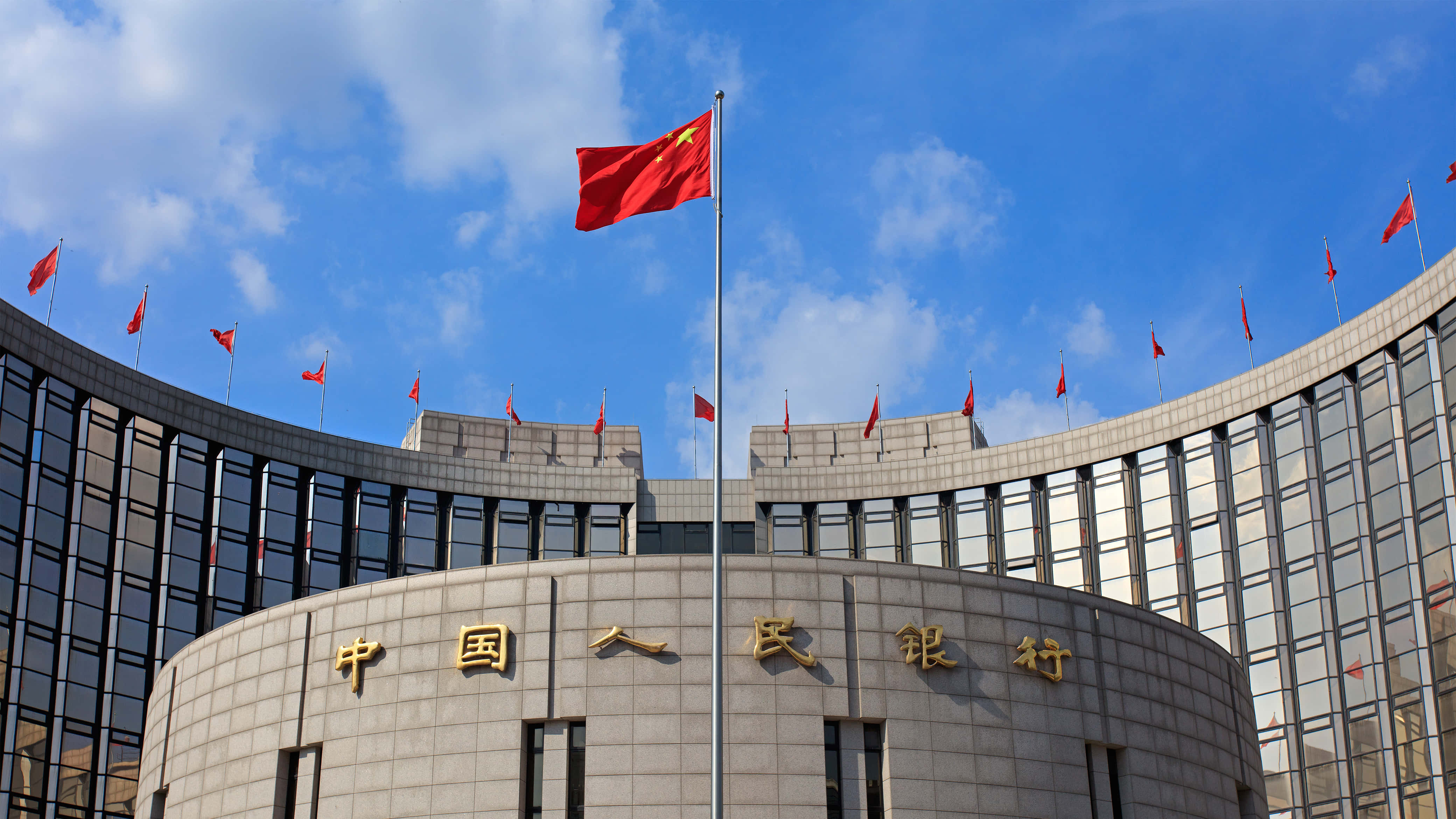 長城日暮時分的迷人風景 壁紙 — 大型弧形建築，上有金色中文字和紅旗，在藍天白雲下飄揚。