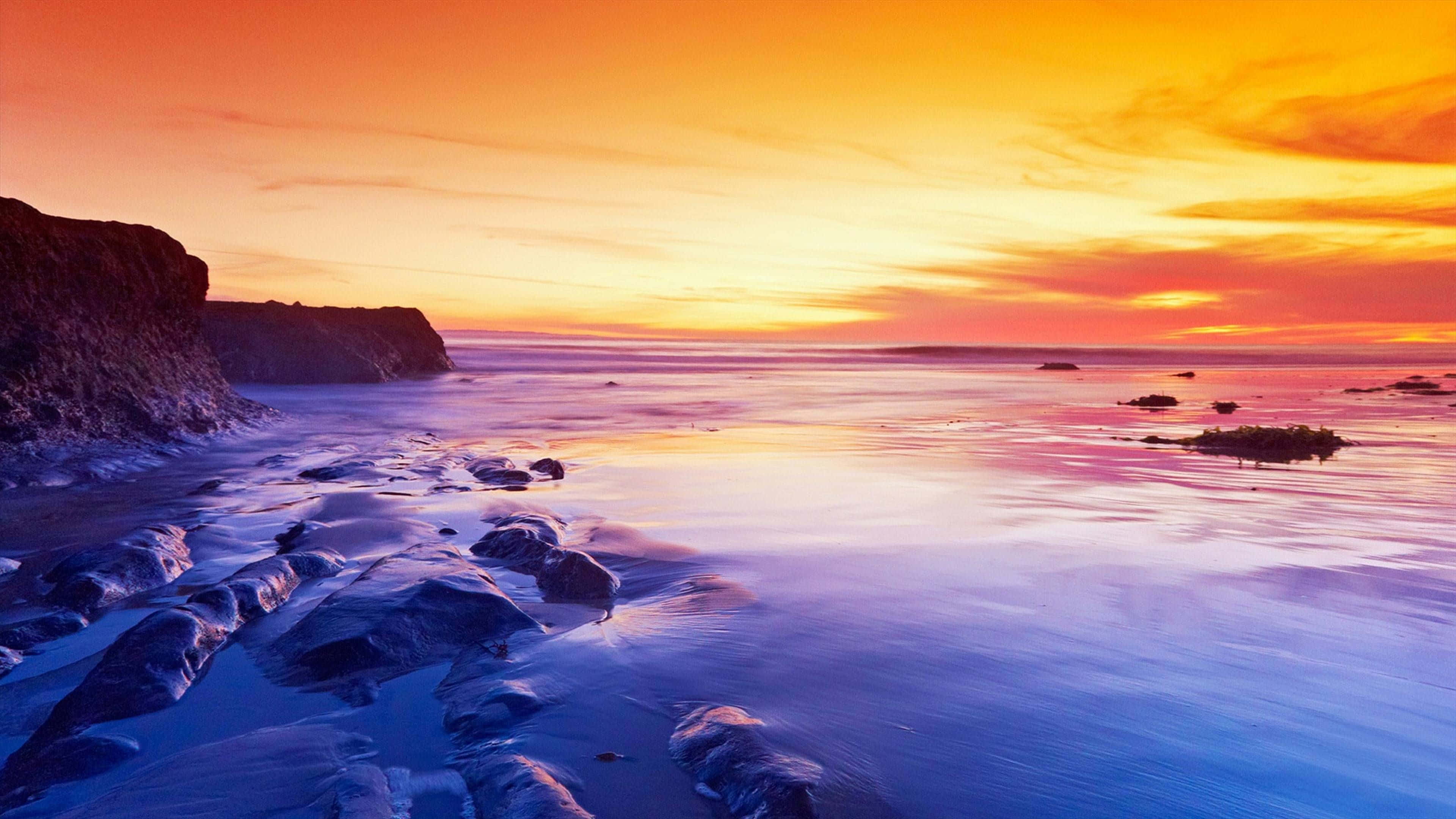 Sunset Photography 桌布 壁紙 — 長時間曝光的日落將海洋染成火熱的橙色和紫色色調，海浪掠過黑暗的沿海岩石。