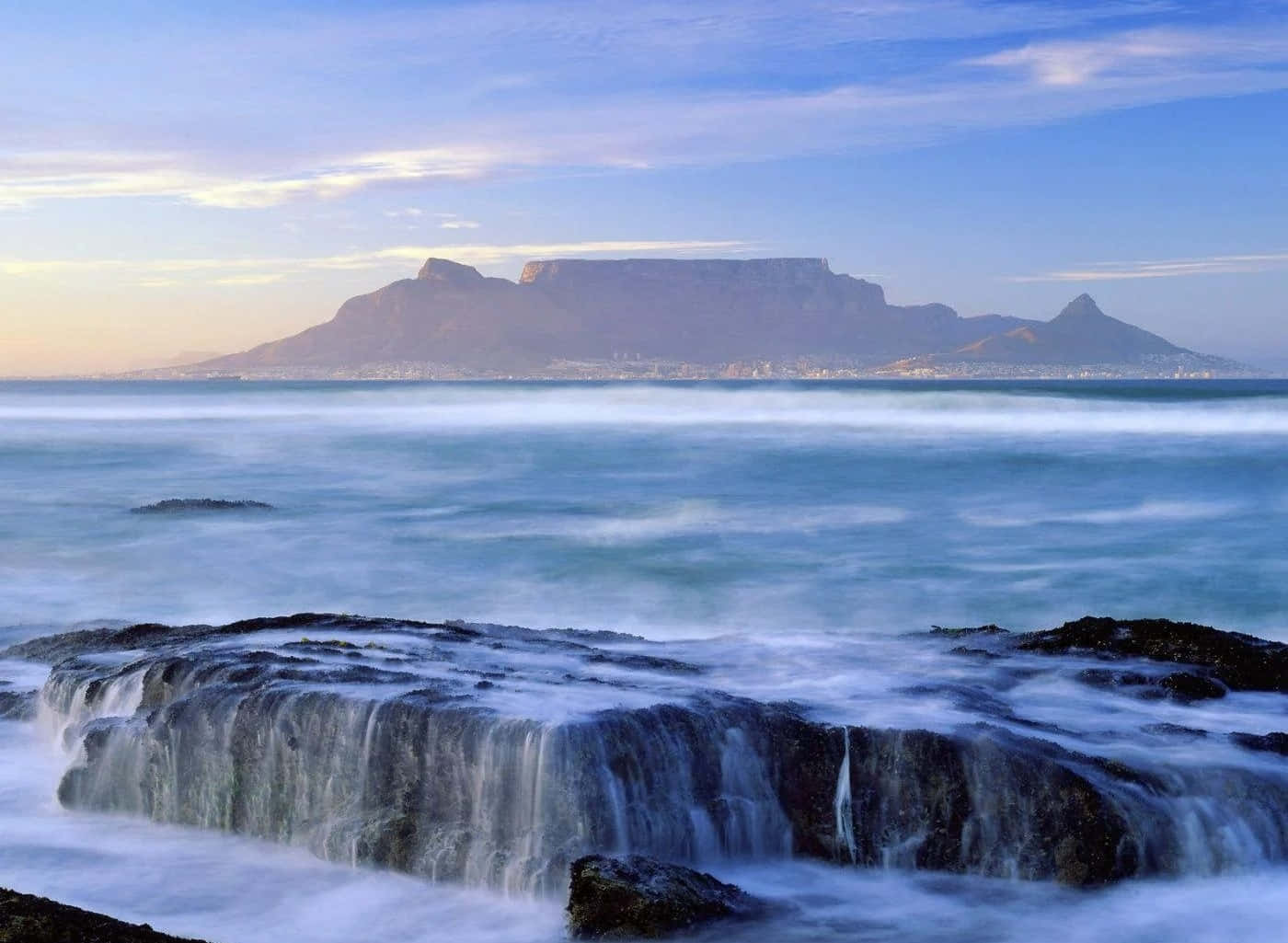 South Africa 桌布 壁紙 — 長時間曝光的海景捕捉到浪花拍打黑色岩石，桌山在柔和的藍天下雄偉地聳立在遠方。