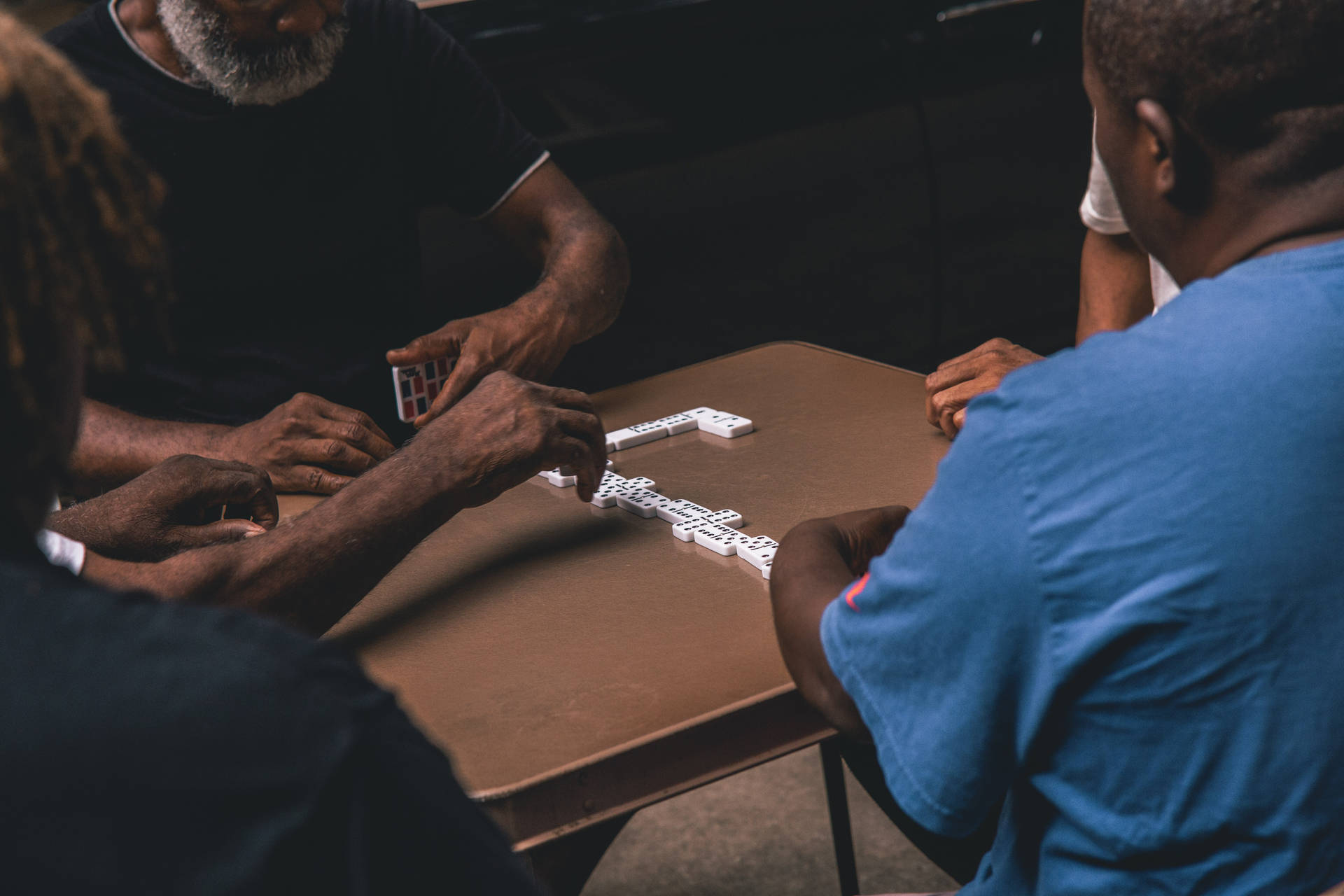 Three men playing dominoes around a wooden table in a dimly lit room.