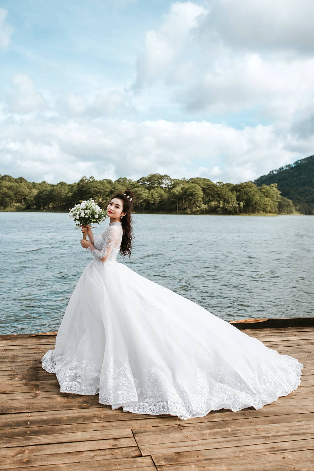 長袖婚紗 壁紙 — 一位新娘穿著寬大的白色蕾絲婚紗，站在平靜湖邊的木製碼頭上，在部分多雲的天空下捧著一束白色鮮花。