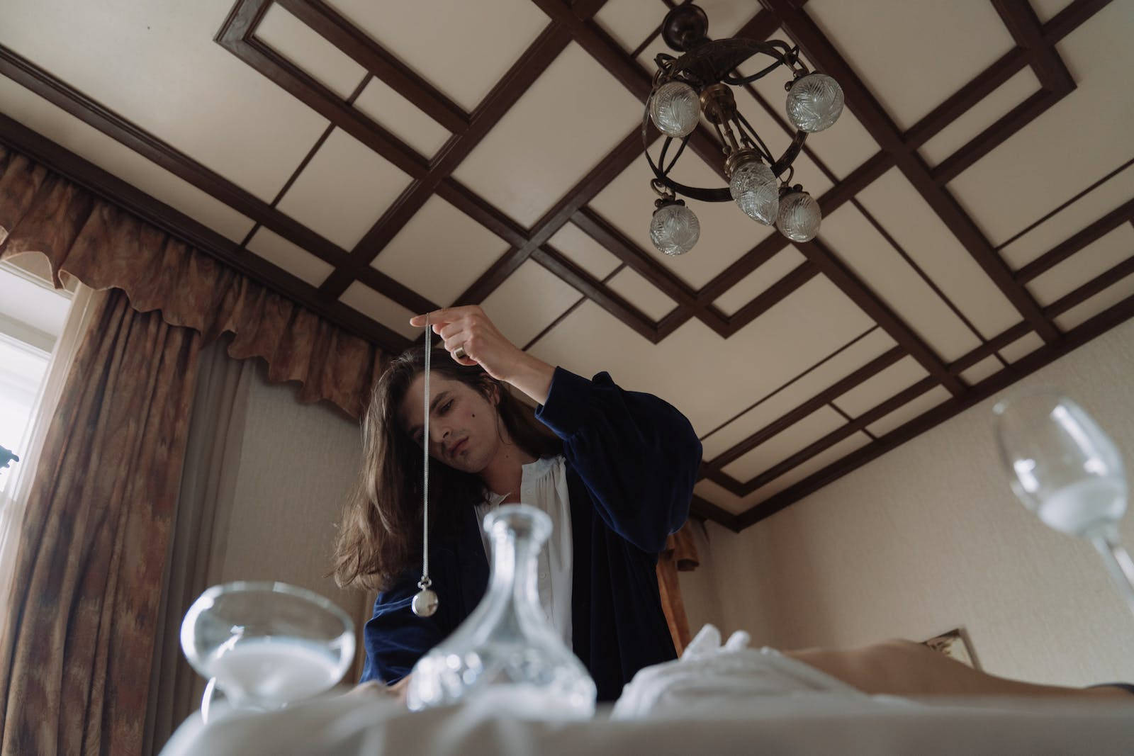 長髮男子進行催眠 壁紙 — 一位身穿藍色長袍的女性在裝飾性天花燈下，手持鐘擺懸於擺放玻璃器皿的桌子上方。