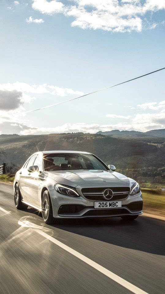 銀色 Mercedes 房車在風景優美的山路上快速行駛，背景有山丘和藍天。