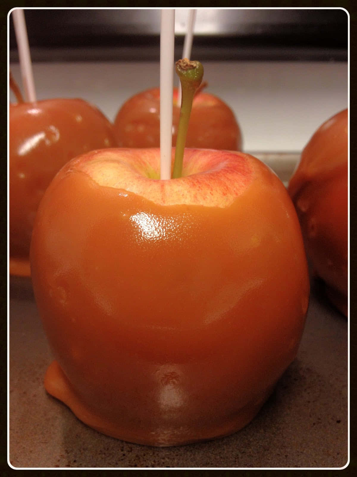 Caramel Apples 桌布 壁紙 — 閃亮的焦糖塗層蘋果插著棍子，放在烤盤上，處於溫暖的室內燈光下。