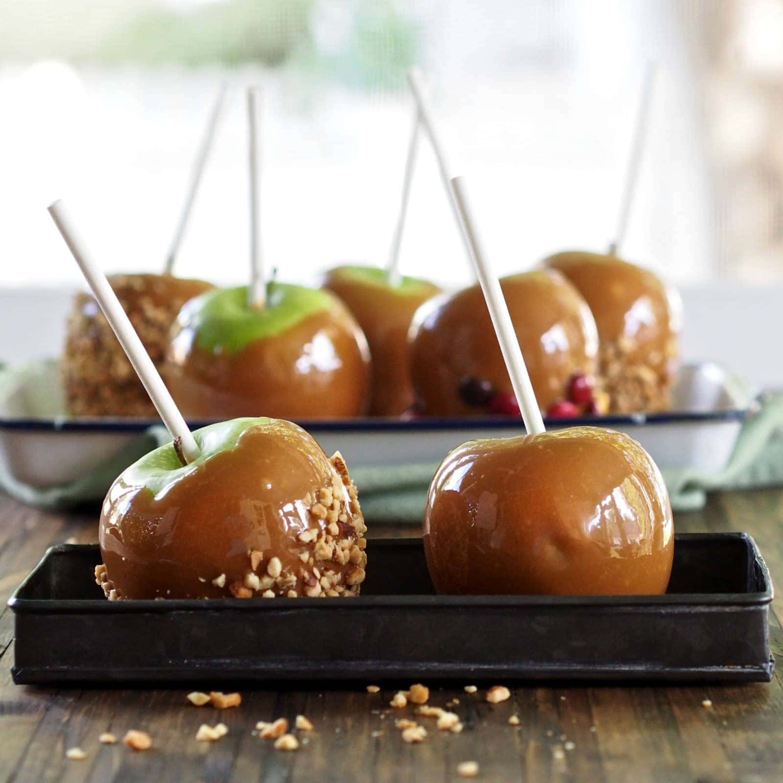 Caramel Apples 桌布 壁紙 — 閃亮的焦糖蘋果，帶有綠色莖和碎堅果，插在棍子上，放在鄉村木桌上的黑色托盤中。