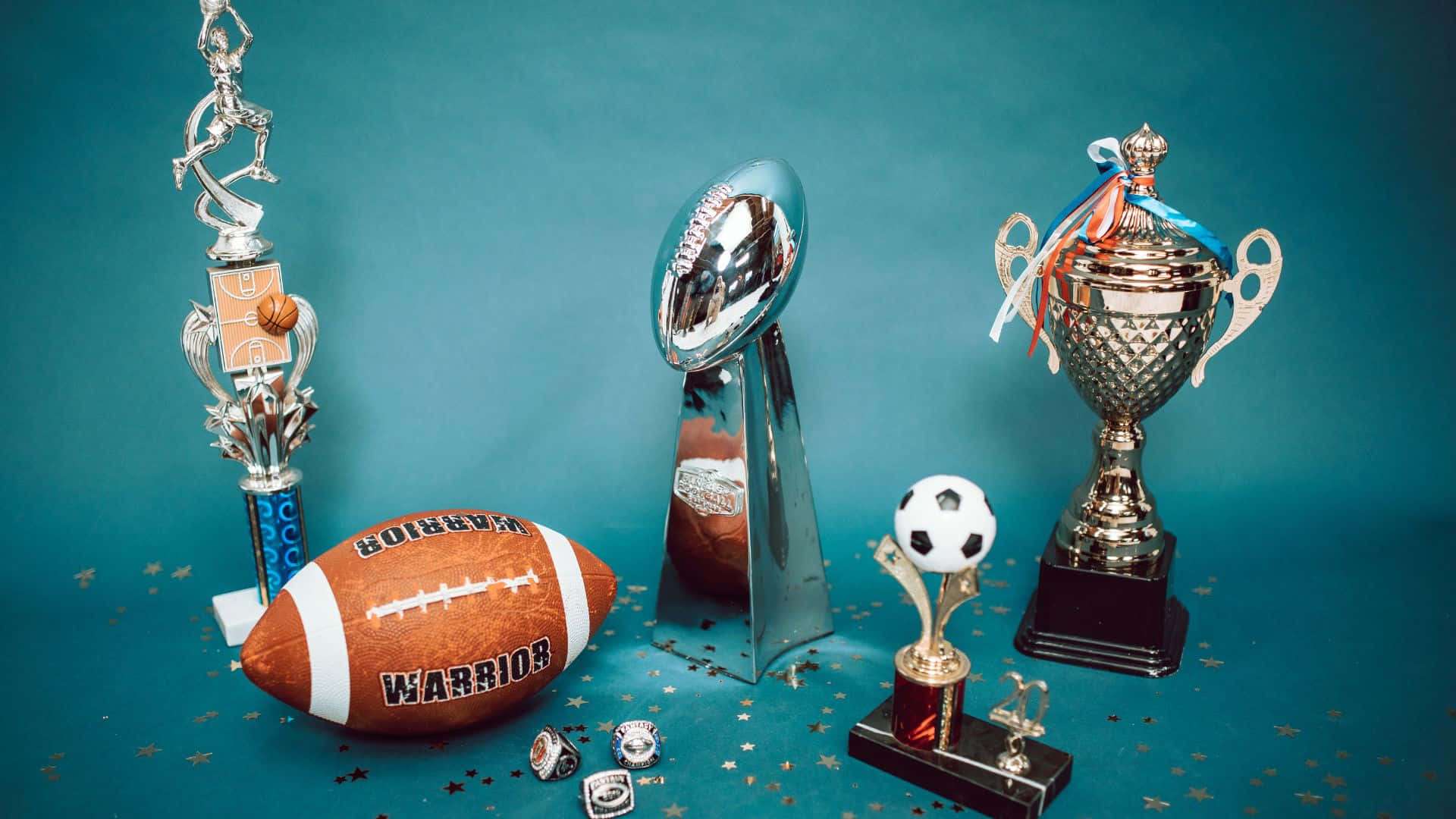 Best Sports 桌布 壁紙 — 閃亮的獎盃和運動球類，如足球和英式足球，排列在帶有閃粉的藍綠色背景上。