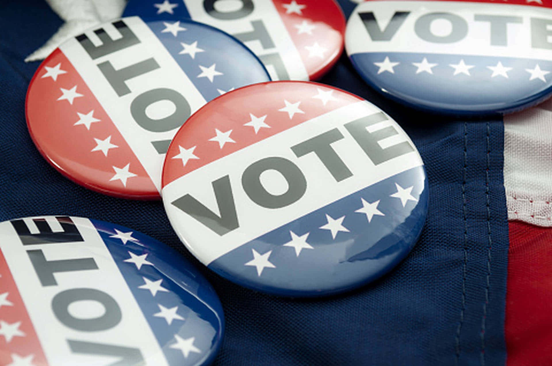 Election 桌布 壁紙 — 閃亮的紅、白、藍投票按鈕帶有星星，放置在帶有紋理的美國國旗布料上。