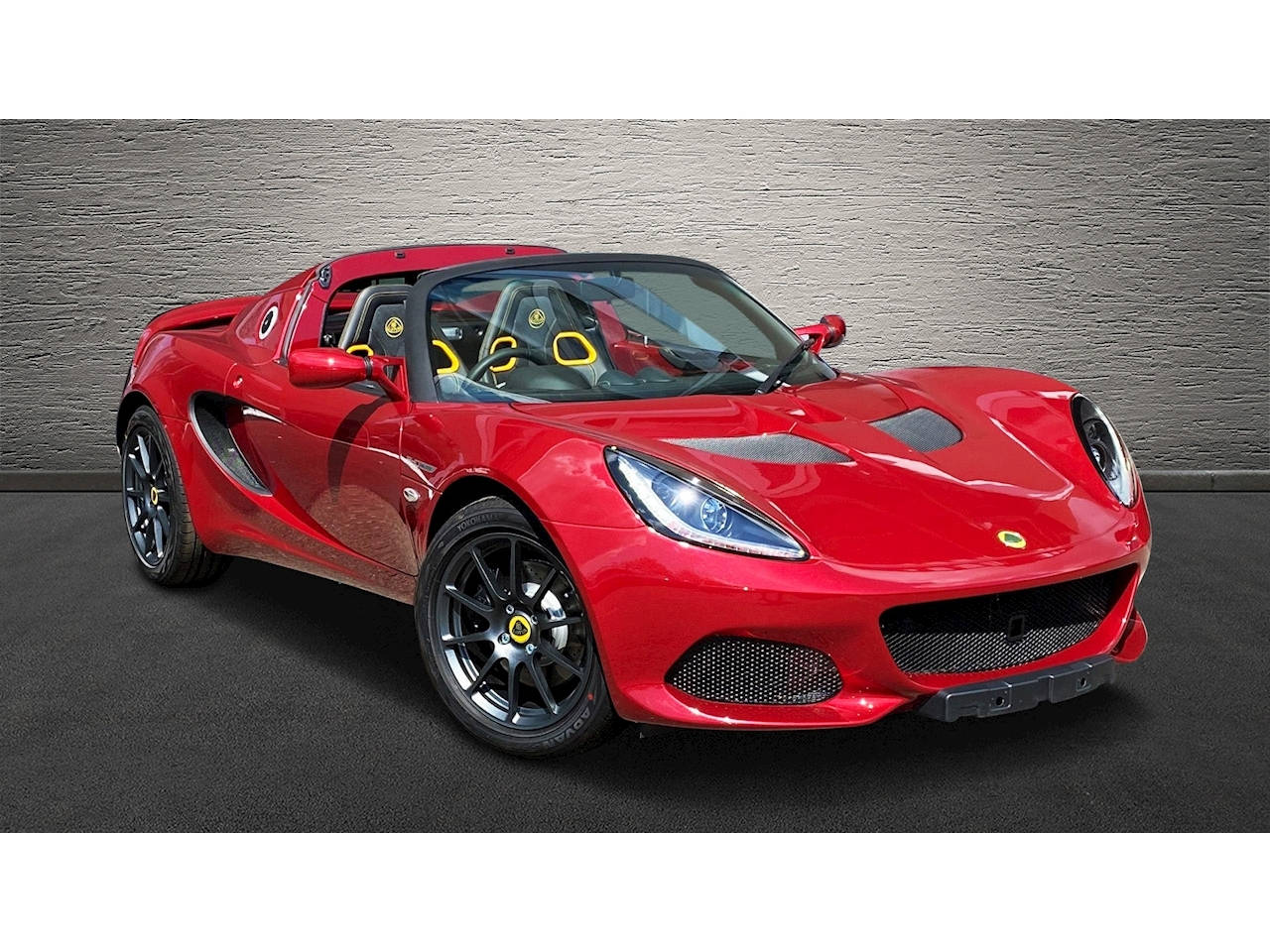 閃亮紅色路特斯Elise跑車 壁紙 — 一輛光澤的紅色蓮花跑車，帶有黑色飾邊和黃色座椅，停靠在有紋理的灰色牆壁前。