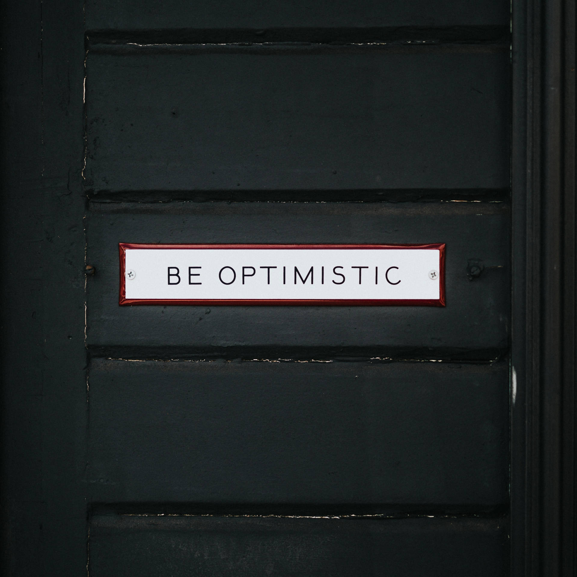 一個白色帶紅色邊框的標誌，上面寫著 BE OPTIMISTIC，位於深綠色木門上。