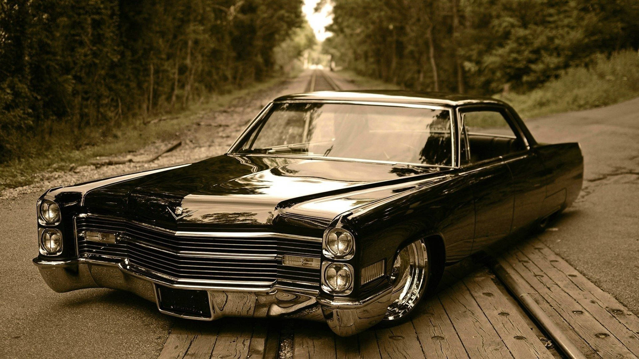 閃耀經典 - 1967年雪佛蘭Impala 壁紙 — 一輛帶有鍍鉻細節的光澤黑色經典汽車停在木板上，旁邊是一條沐浴在溫暖懷舊色調光線中的森林道路。