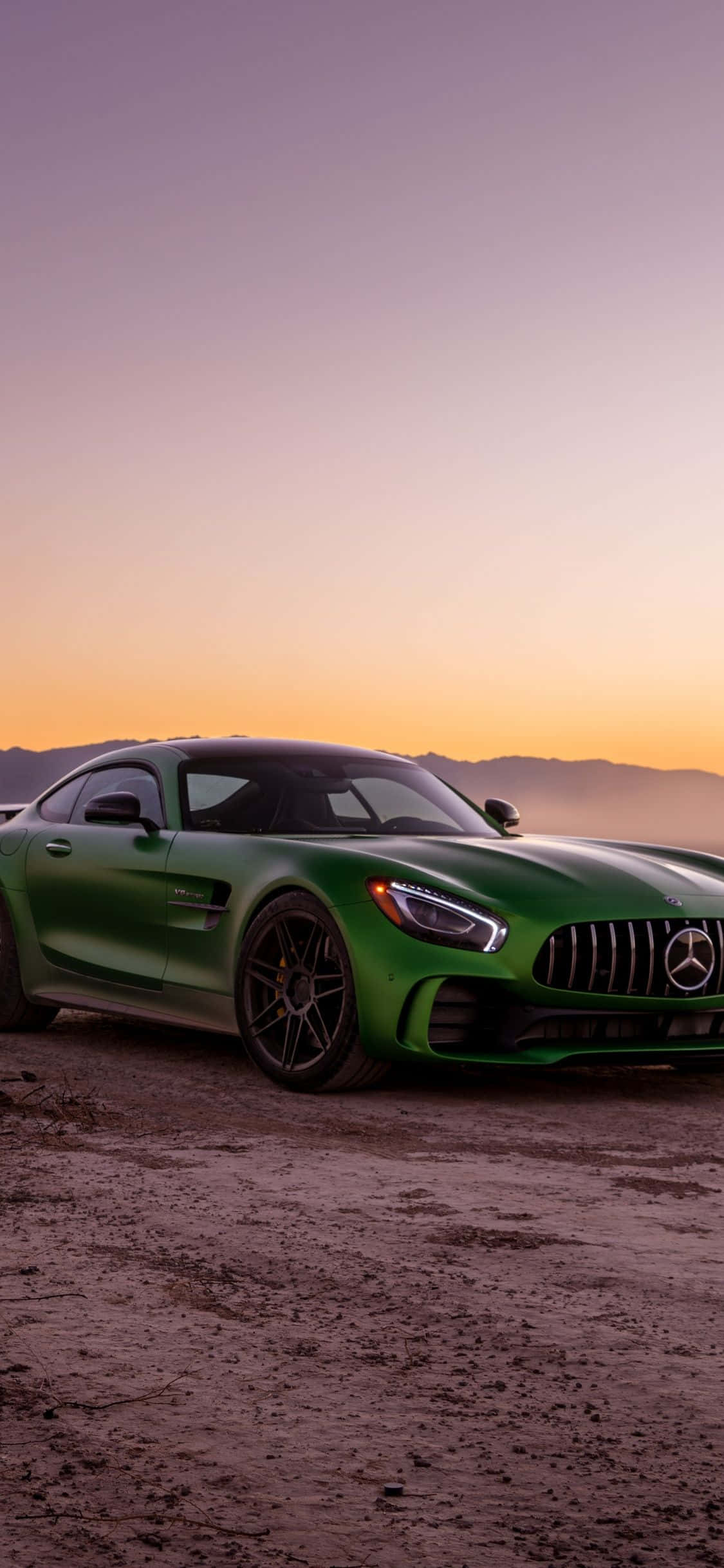 閃耀綠色iPhone XS與梅賽德斯-AMG GT山頂日落背景 壁紙 — 一架光澤的綠色 Mercedes-AMG GT 停泊在塵土飛揚的地形上，背景是 vibrant 的日落天空和遠處的山脈。