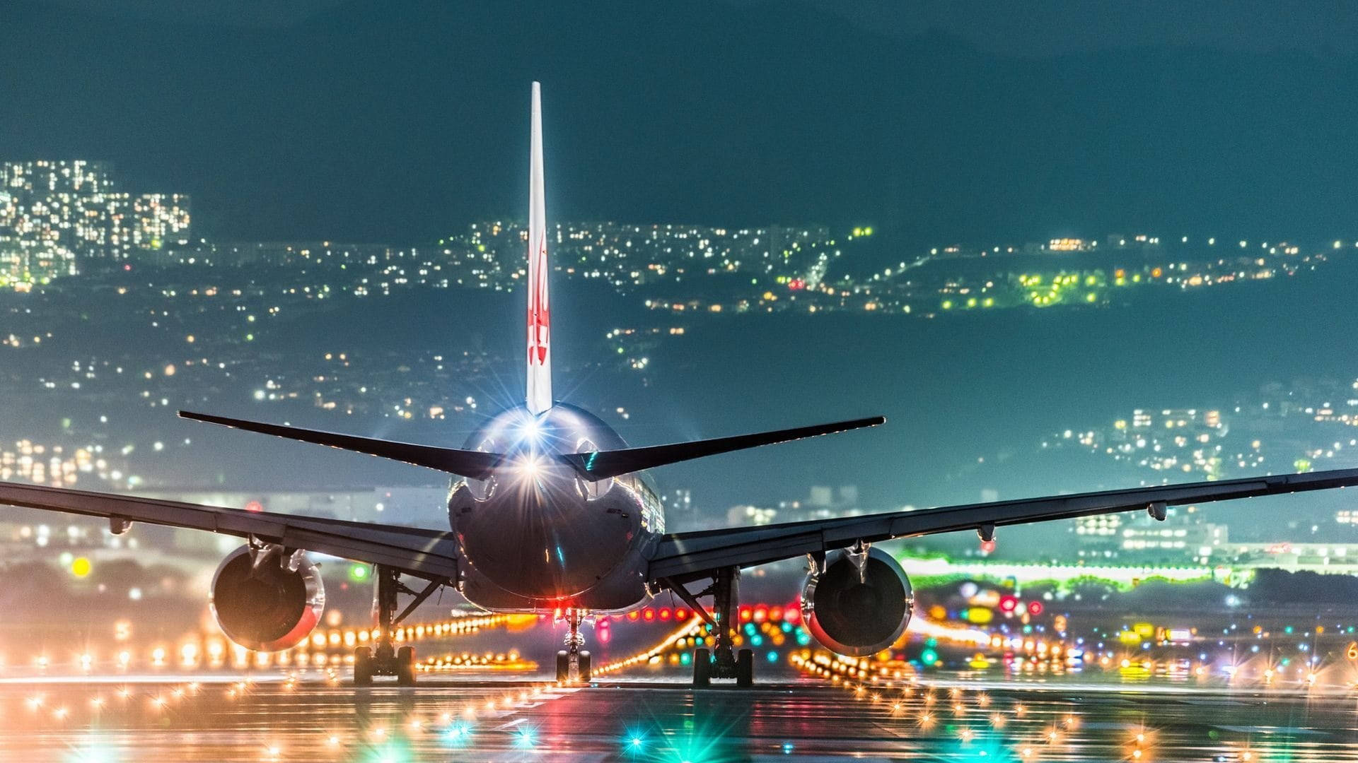 閃耀跑道城市夜景 壁紙 — 一架大型飛機在夜間停在濕滑的跑道上，背景中城市燈光閃爍。