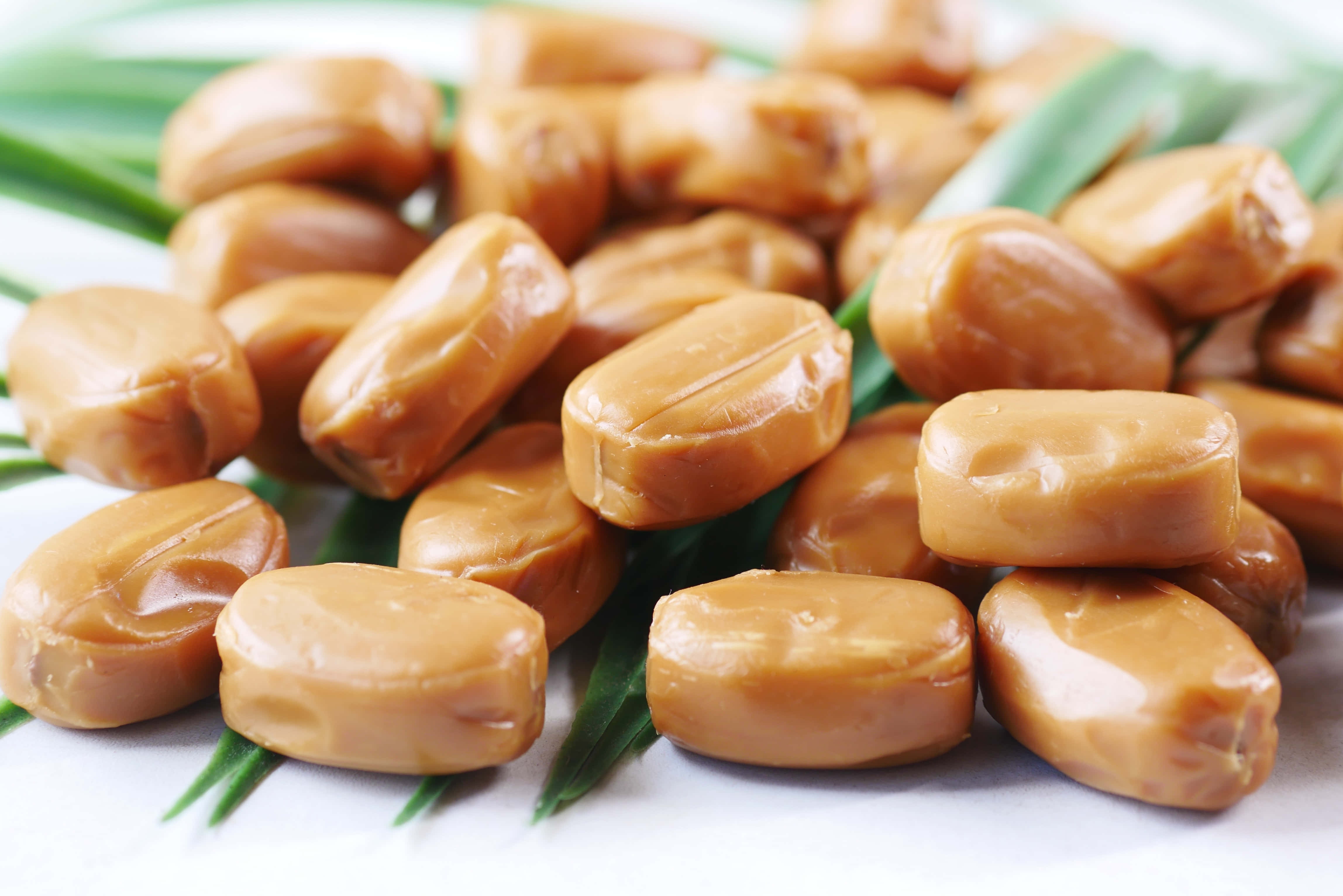 Caramel 桌布 壁紙 — 閃閃發光的金色焦糖糖果堆在綠色棕櫚葉上，背景是明亮的白色。