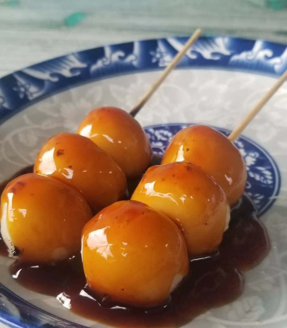 Japanese Sweets 桌布 壁紙 — 閃閃發光的金黃色餃子串放在深色醬汁中，置於藍白圖案碟上。