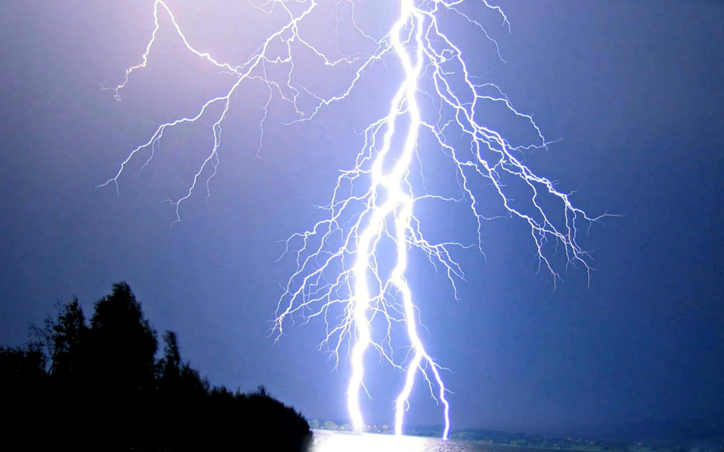 閃電之擊 壁紙 — 一道巨大的叉狀閃電照亮了暗藍色的夜空，下方是樹木的剪影和反射的水面。