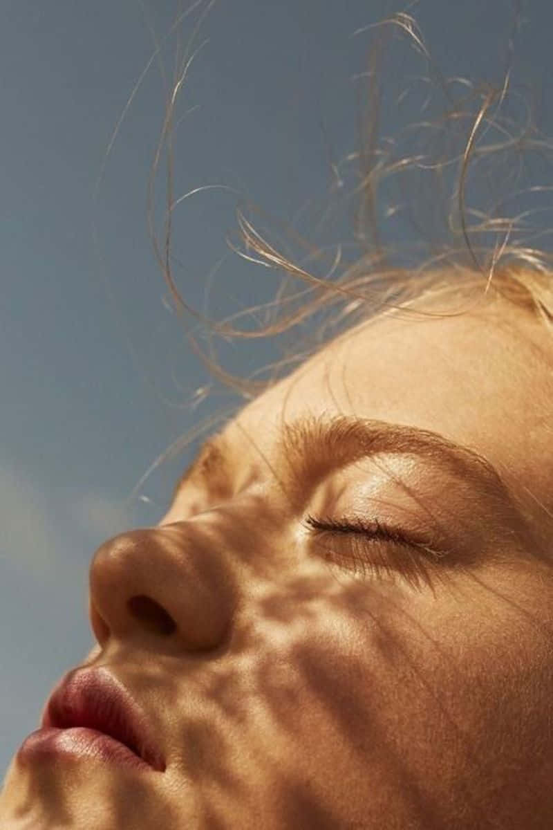閉眼沐浴陽光的女孩 壁紙 — 一個人閉上眼睛的臉部特寫，沐浴在溫暖的陽光中，陽光在皮膚上投下樹葉的影子，背景是清澈的藍天。