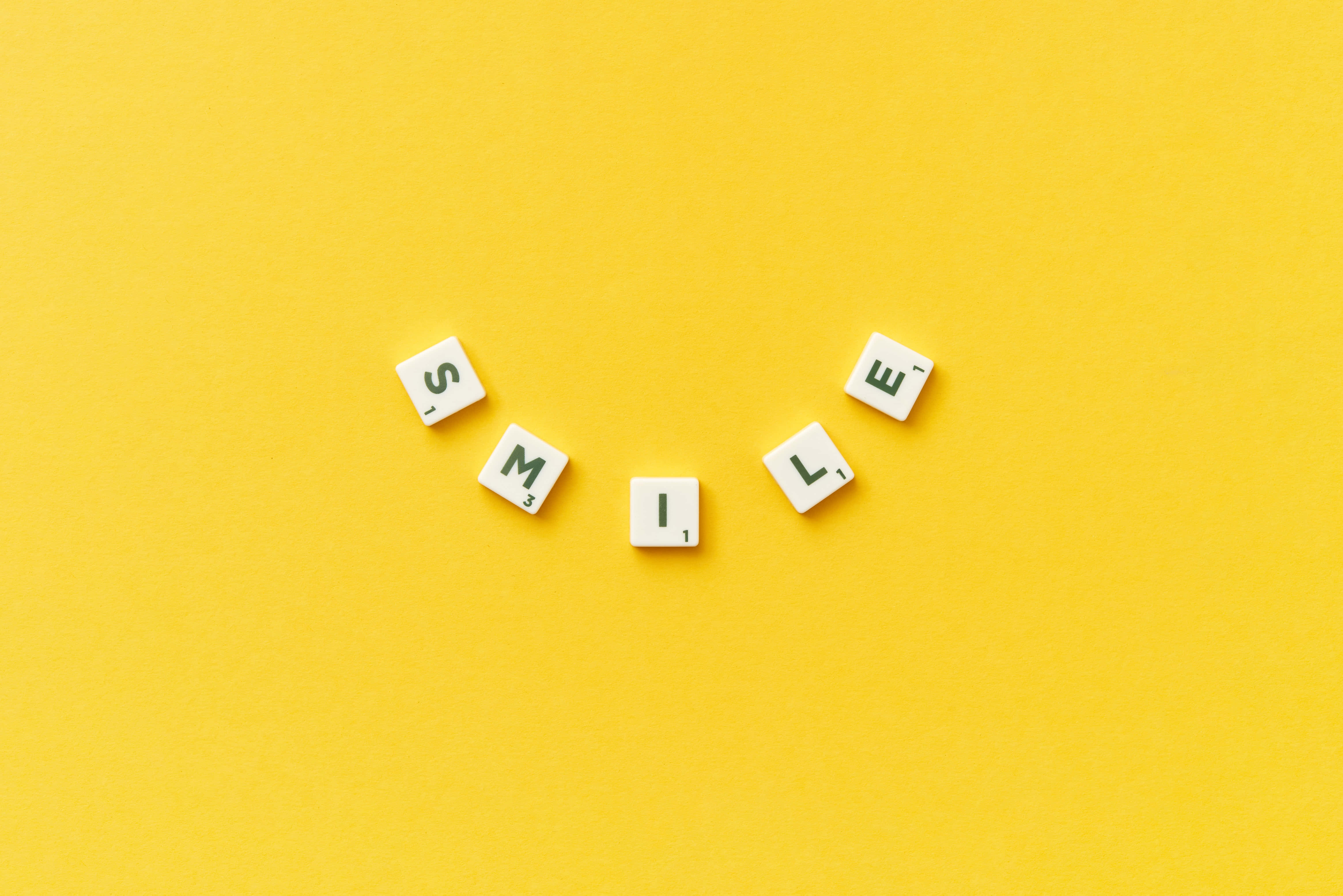 開始微笑吧 壁紙 — 白色字母瓷磚拼出 'SMILE'，以彎曲微笑形狀排列在鮮豔的黃色背景上。