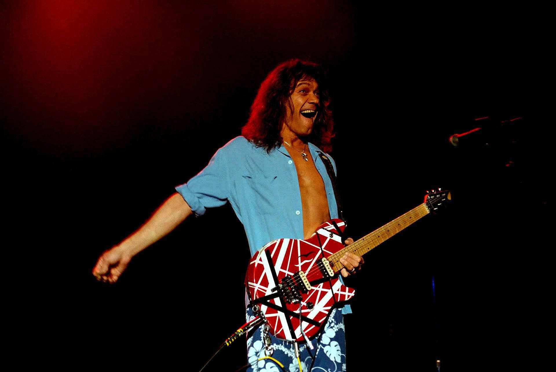 開心愛迪·范·海倫演出 壁紙 — Eddie Van Halen 在紅色燈光下於舞台上張開雙臂，帶著喜悅的表情彈奏結他。