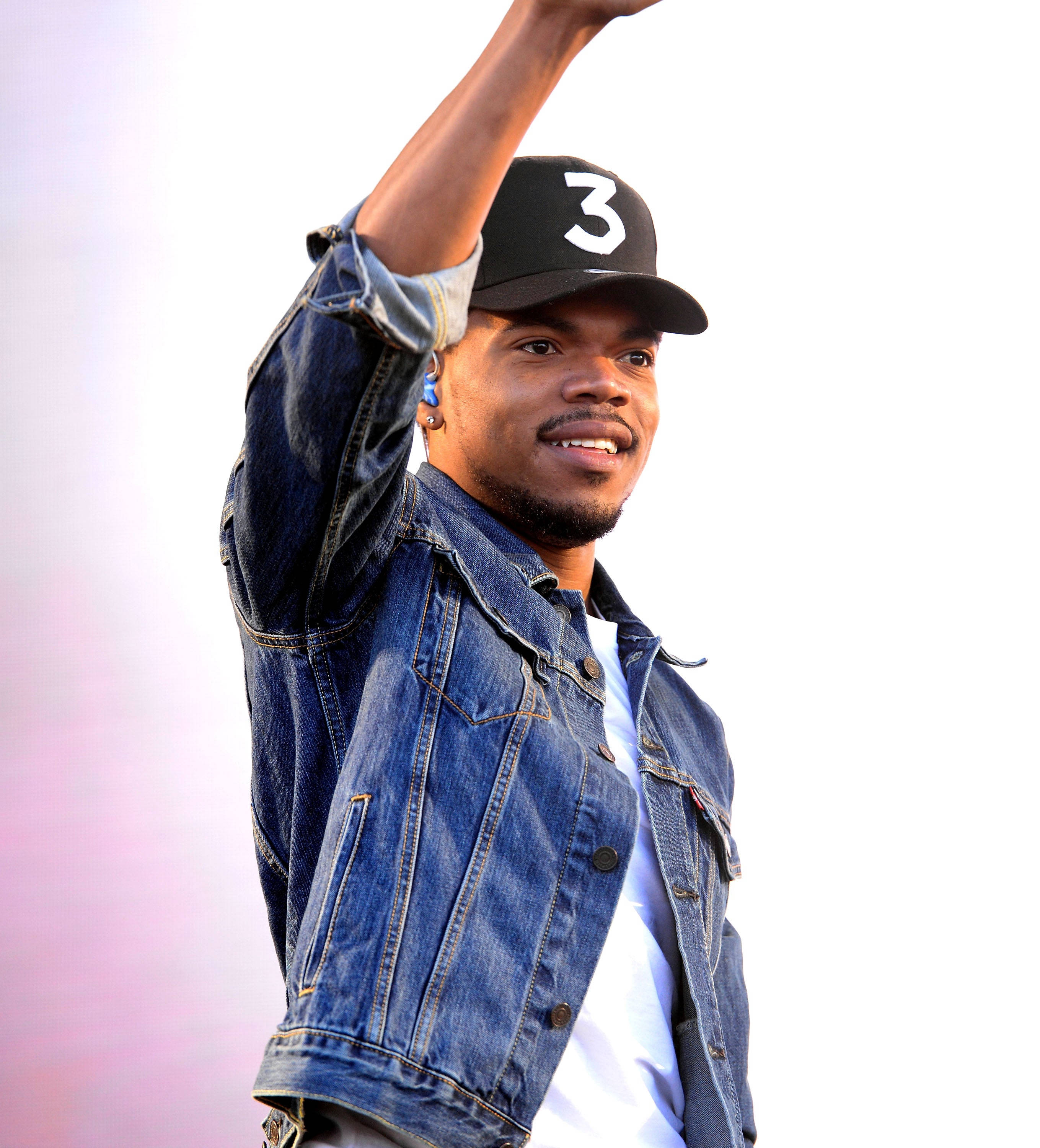 開心的Chance the Rapper 壁紙 — 一個微笑的男人穿著牛仔外套和黑色帽子，帽上有白色數字三，在柔和的粉紅色和白色背景前舉起手臂。
