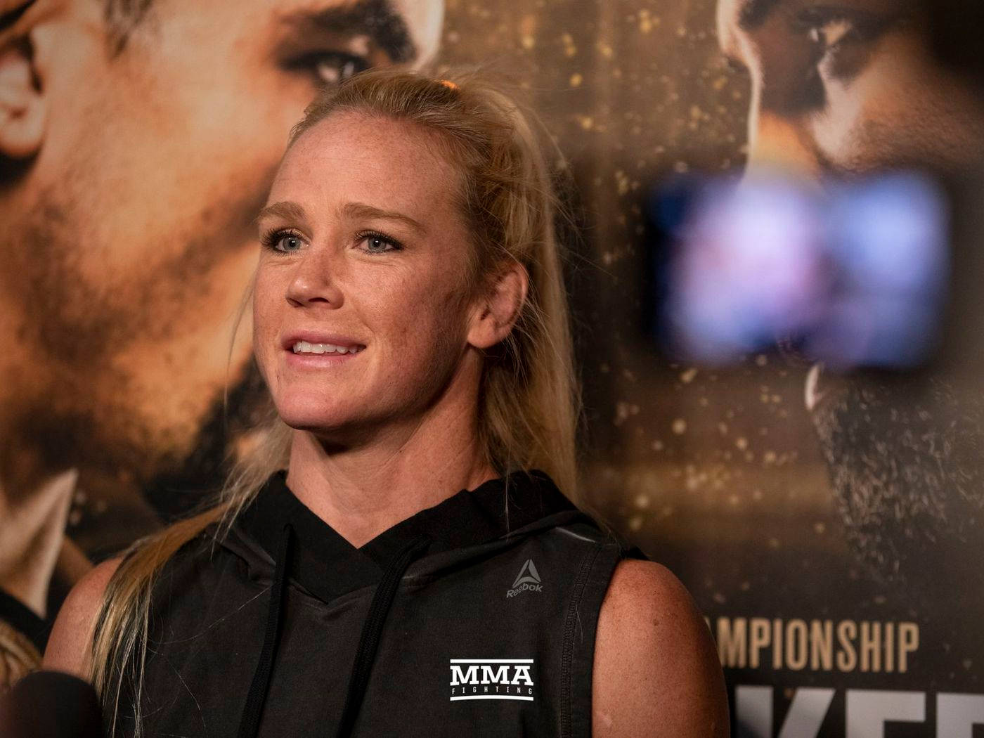 開心的Holly Holm 壁紙 — 一位身穿黑色無袖連帽衫的女性MMA拳手在記者會上溫柔微笑，身後有冠軍海報。