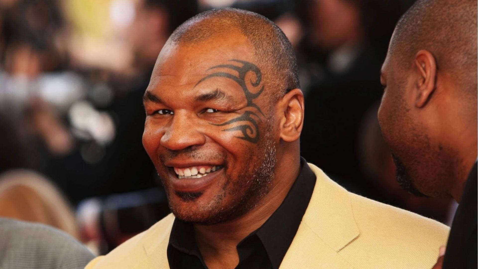 開心的邁克·泰森 壁紙 — Mike Tyson 露出燦爛笑容，臉頰上可見其標誌性的部落紋身，身穿米色西裝。