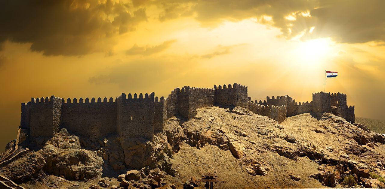開羅古堡 Citadel 壁紙 — 古老石堡壘坐落在岩石山頂上，沐浴在金色的日落光芒中，頂部有旗幟飄揚。