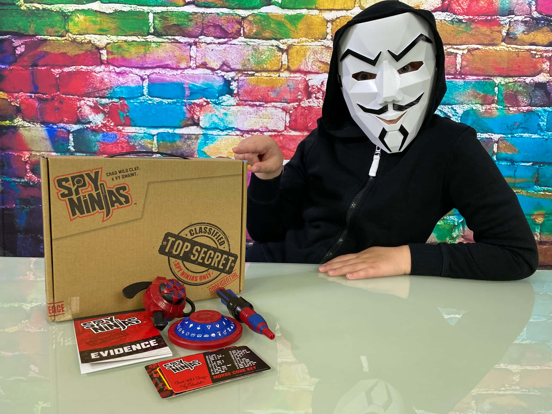 戴面具嘅細路喺枱上展示 Spy Ninjas 玩具，背景係色彩繽紛嘅磚牆。