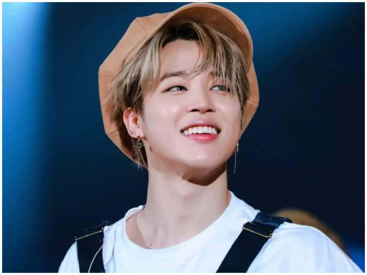 年輕男子戴著棕褐色帽子、穿著白襯衫，在舞台燈光下微笑。 — Jimin GIF 壁紙