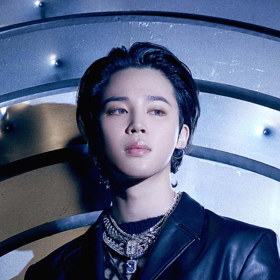 防彈少年團Jimin身穿色彩繽紛服裝的時尚造型 壁紙 — 一位深色頭髮、佩戴華麗珠寶的年輕男子，在冷藍色燈光下，抬頭望向彎曲的金屬背景。