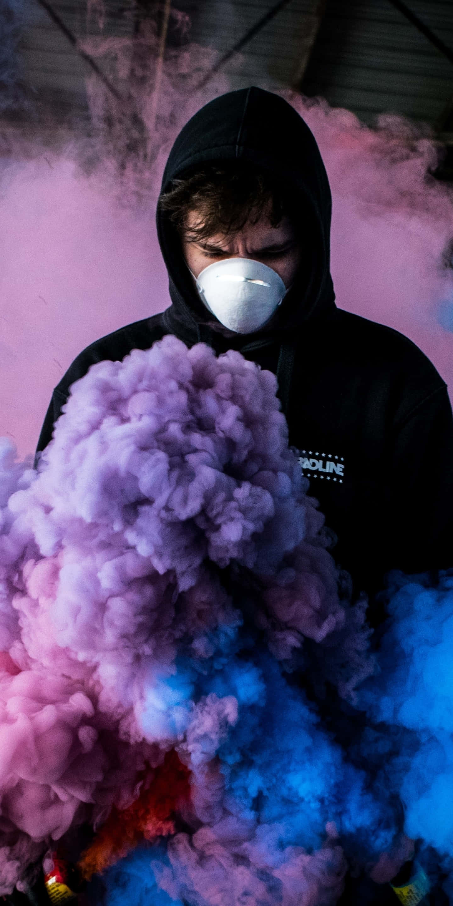 防毒面具男子圖片 壁紙 — 一個穿著黑色連帽衫和白色口罩的人站在工業環境中，周圍環繞著旋轉的粉紅色和藍色煙霧。