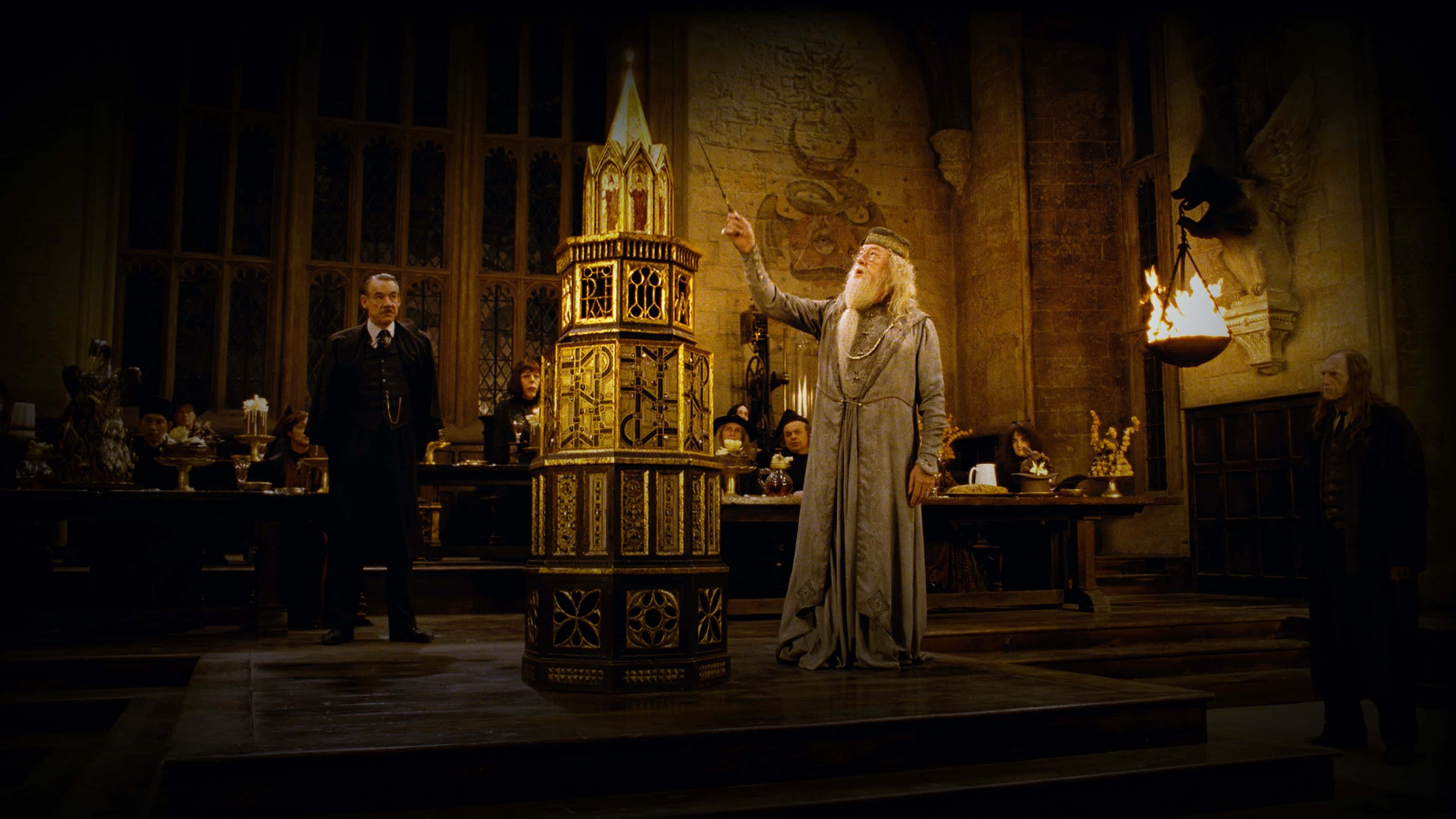 The Goblet Of Fire 桌布 壁紙 — 阿不思·鄧不利多站在一個宏偉、燈光昏暗的大廳中，揮舞魔杖指向一座高聳華麗的金色結構，其他人則莊嚴地注視著。