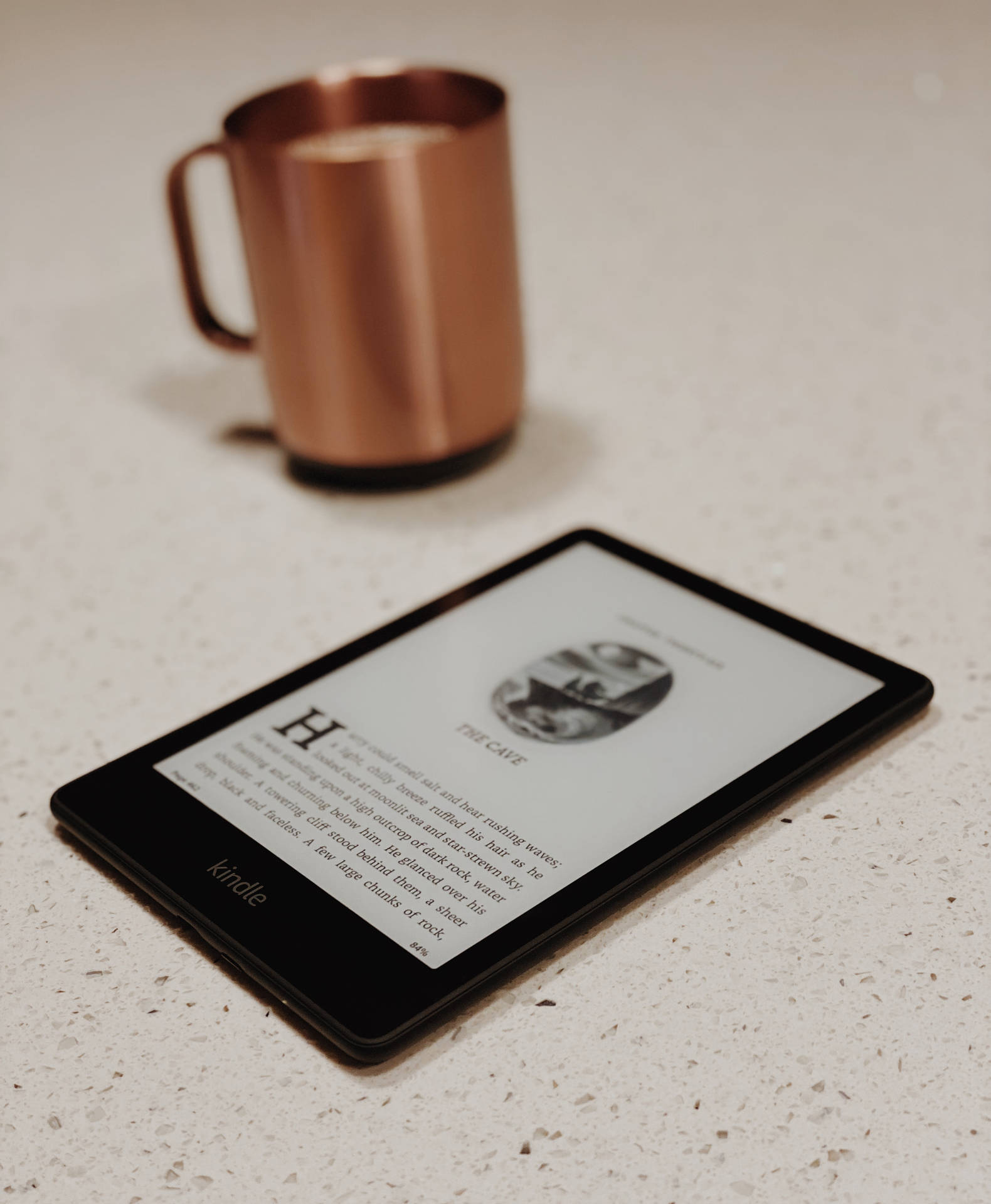 Kindle顯示書頁，旁邊有銅杯，放在有斑點的檯面上。