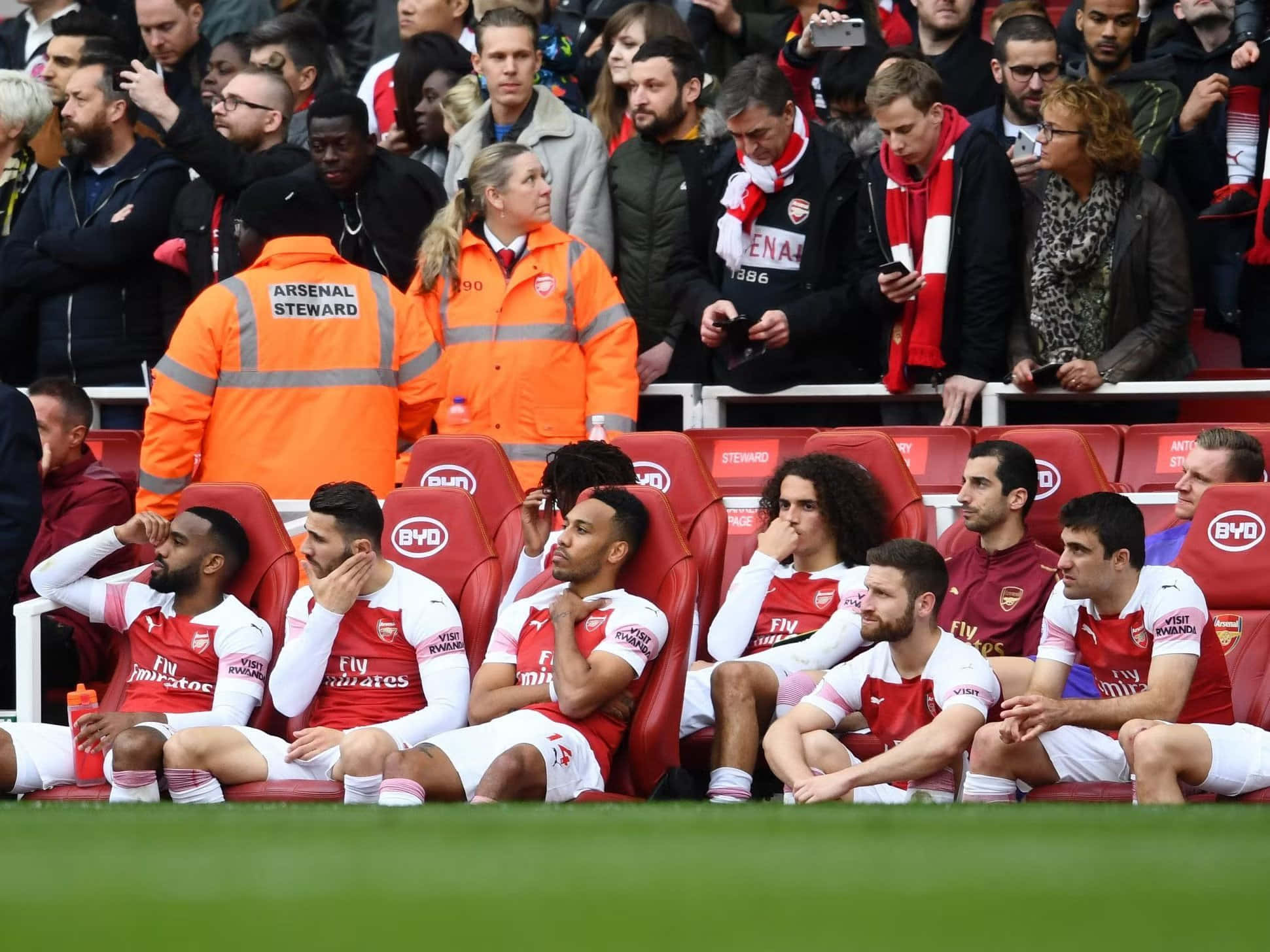 Arsenal 桌布 壁紙 — 阿仙奴足球員身穿紅白球衣沮喪地坐在後備席上，身穿橙色外套的工作人員站在他們身後。