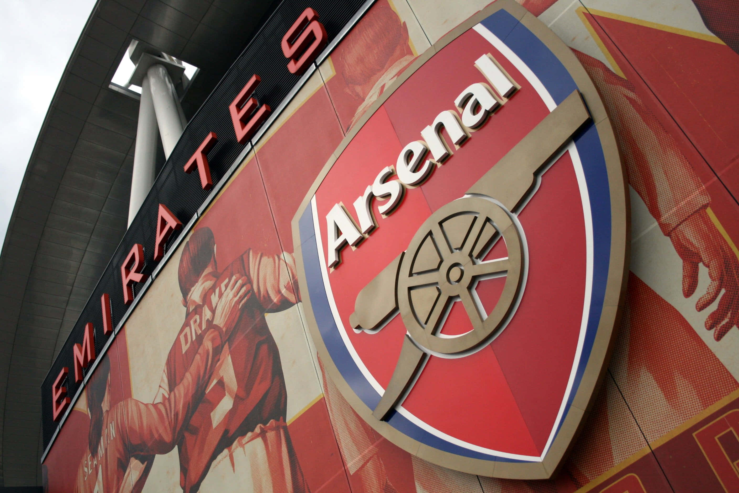 Arsenal 桌布 壁紙 — 阿仙奴足球會標誌性徽章在酋長球場外牆的特寫，旁邊有球員壁畫。