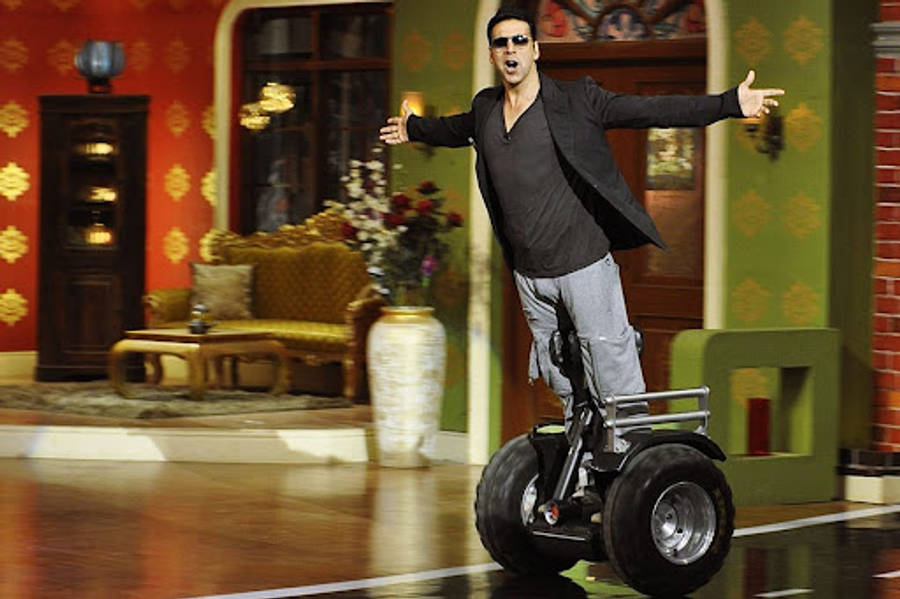 阿克謝·庫馬爾騎Segway電動車 壁紙 — 一名戴著太陽眼鏡、穿著休閒服裝的男子，在裝飾華麗的電視攝影棚場景中，騎著兩輪電動滑板車保持平衡。