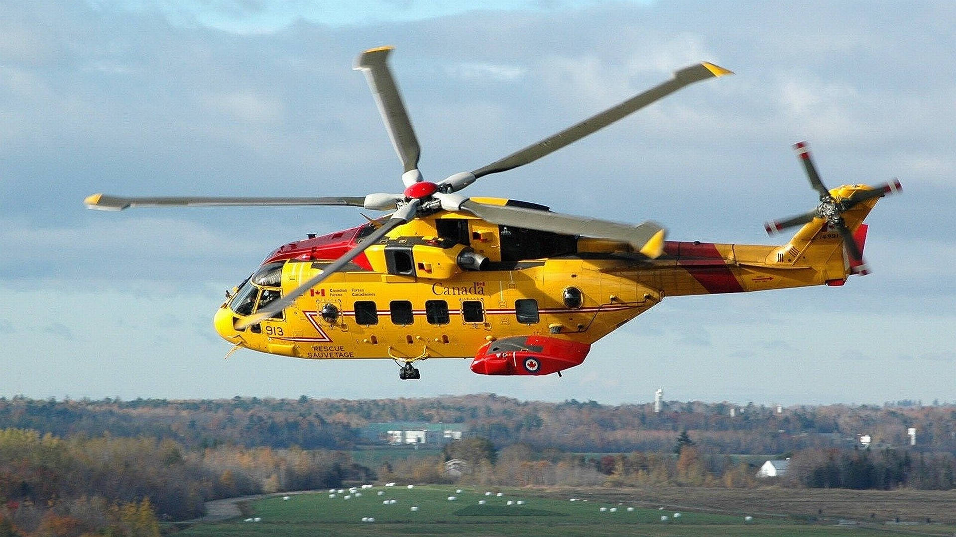 阿古斯塔韋斯特蘭 CH-149 直升機 4K 壁紙 — 一架亮黃色的加拿大搜索救援直升機在陰天低空飛過鄉村景觀，有秋天的樹木和綠色的田野。