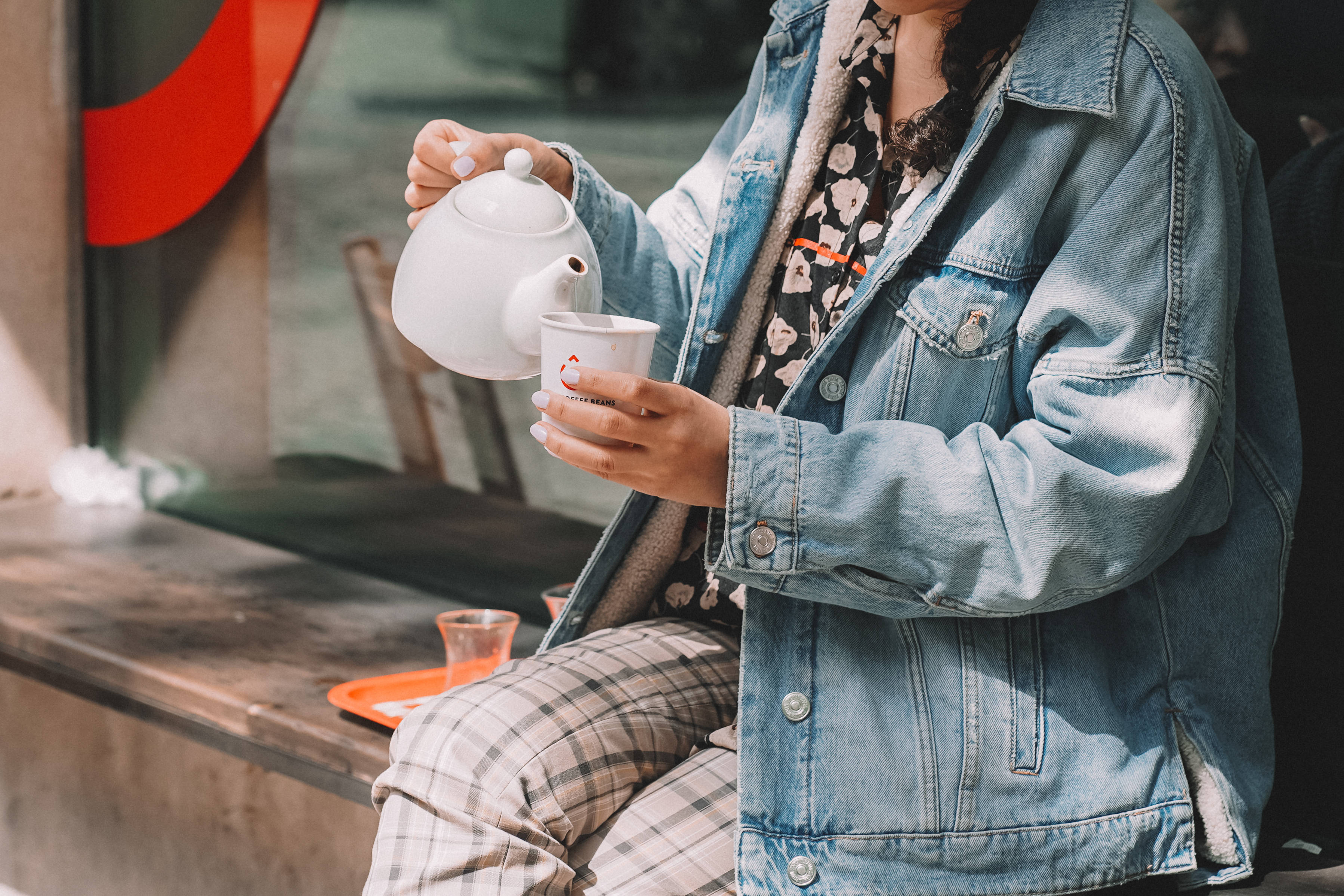 阿塞拜疆品茶時光 壁紙 — 一位穿著牛仔外套的人坐在木長椅上，將白色水壺中的茶倒入杯中。